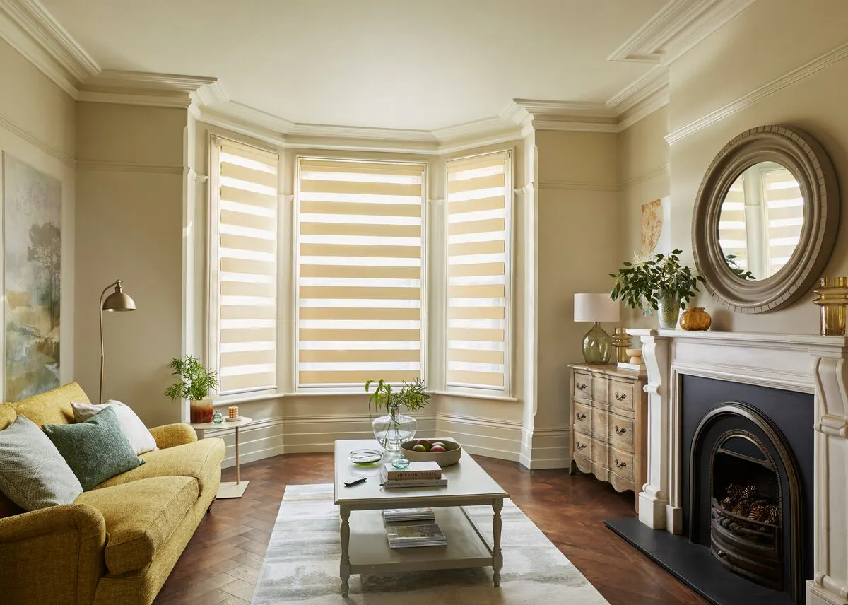 Cosy living room with bay windows fitted with striped day and night blinds, a yellow sofa, fireplace, and a central coffee table.