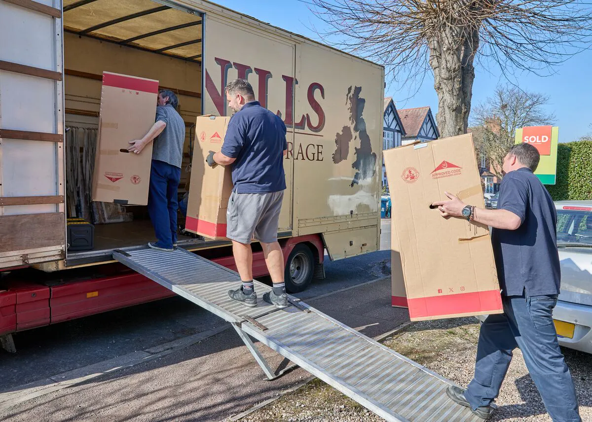 People loading large cardboard boxes into a removals truck using a metal ramp on a sunny moving day.