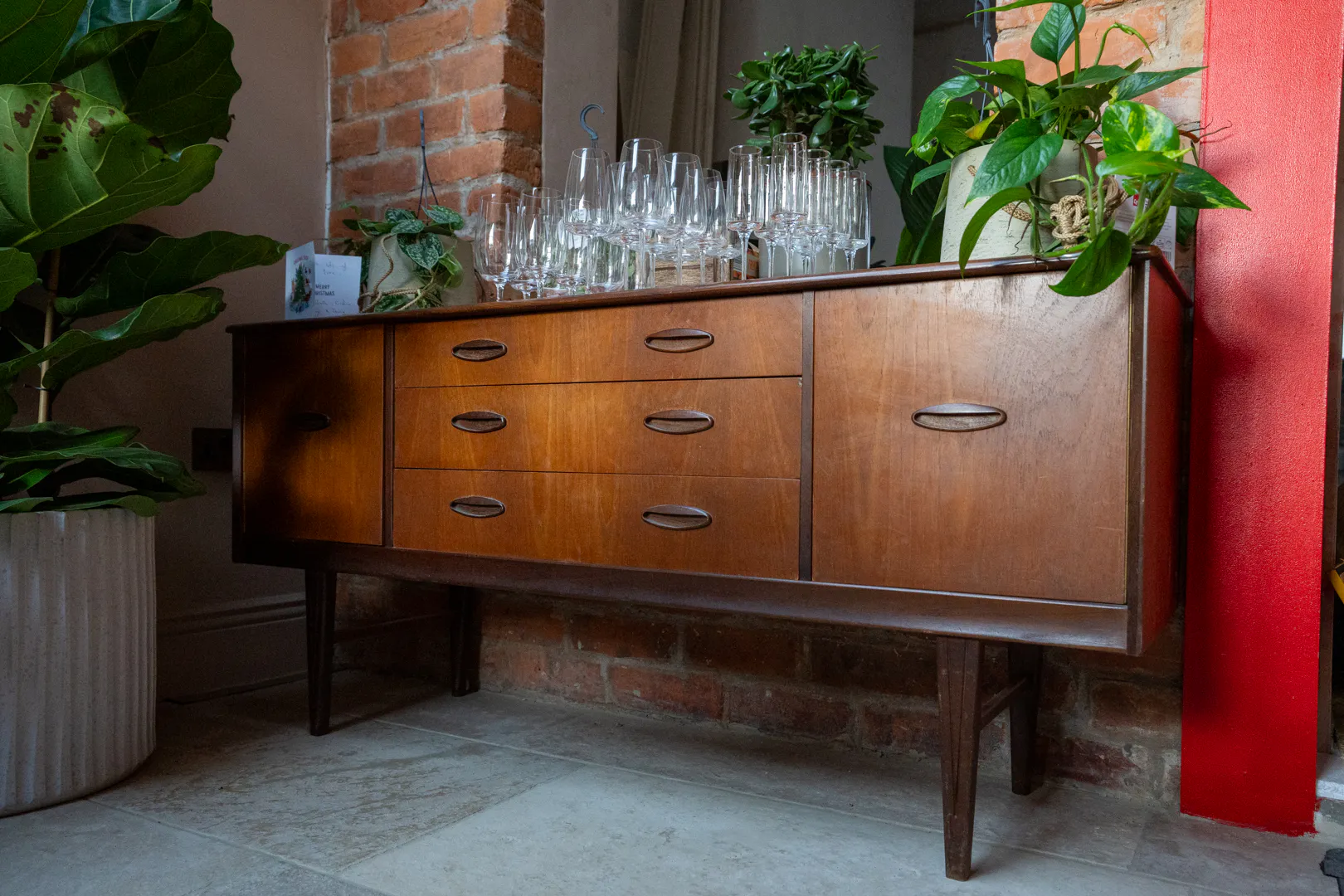 Mid-century wooden sideboard with glassware and potted plants displayed on top, set against exposed brick and a red accent wall.