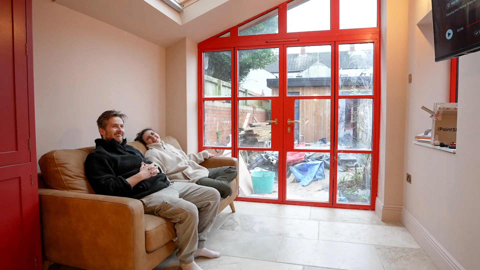 Two people relaxing on a sofa in a bright room with red-framed glass doors overlooking a cluttered garden area.