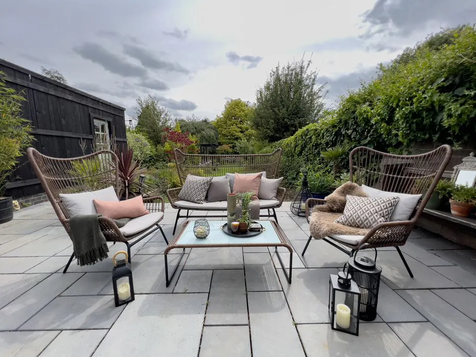 Outdoor patio with wicker chairs, a sofa, cushions and lanterns arranged around a glass-top table in a leafy garden.