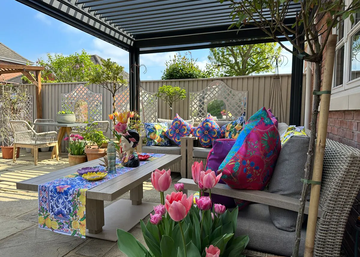 Colourful outdoor seating area with patterned cushions, tulips and plants under a pergola in a sunny garden.