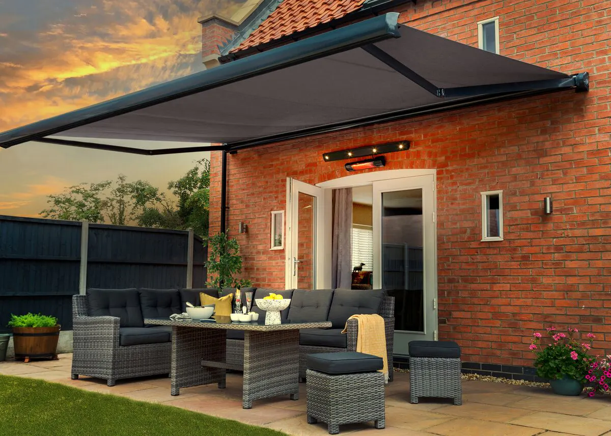 Patio with grey rattan furniture beneath a large retractable awning, set against a brick house at sunset.