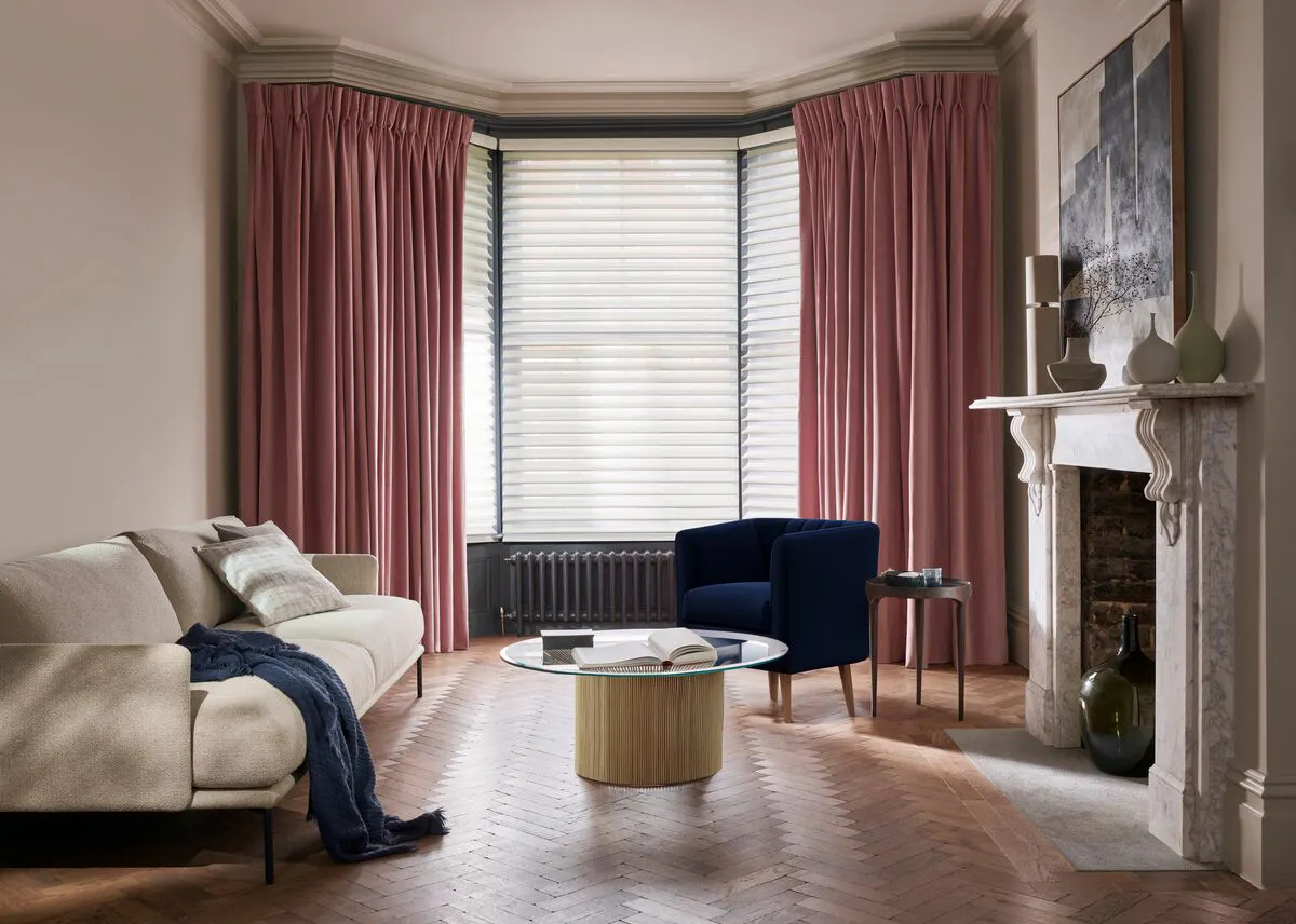 Elegant living room with pink curtains, a beige sofa, navy armchair and glass coffee table beside a marble fireplace.