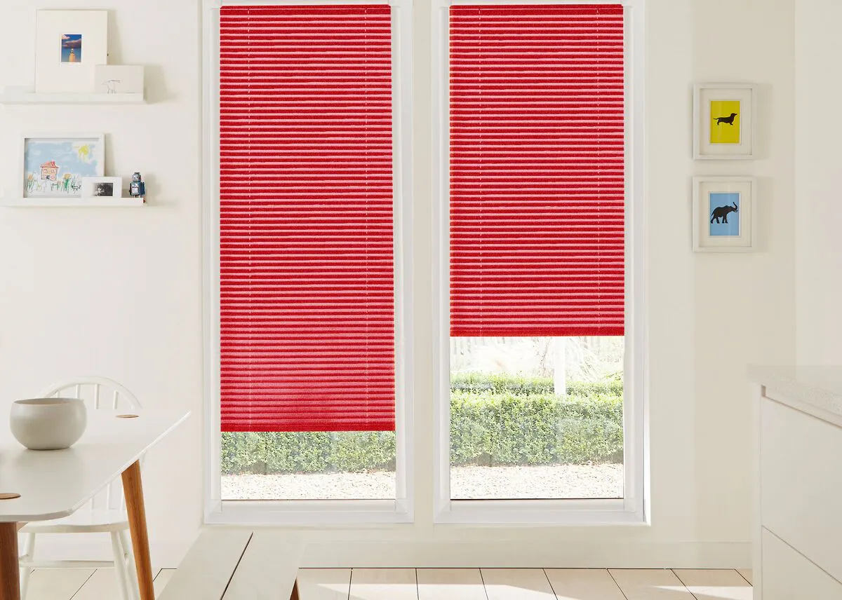Bright kitchen with two large windows fitted with vibrant red pleated blinds, overlooking a neat green hedge.