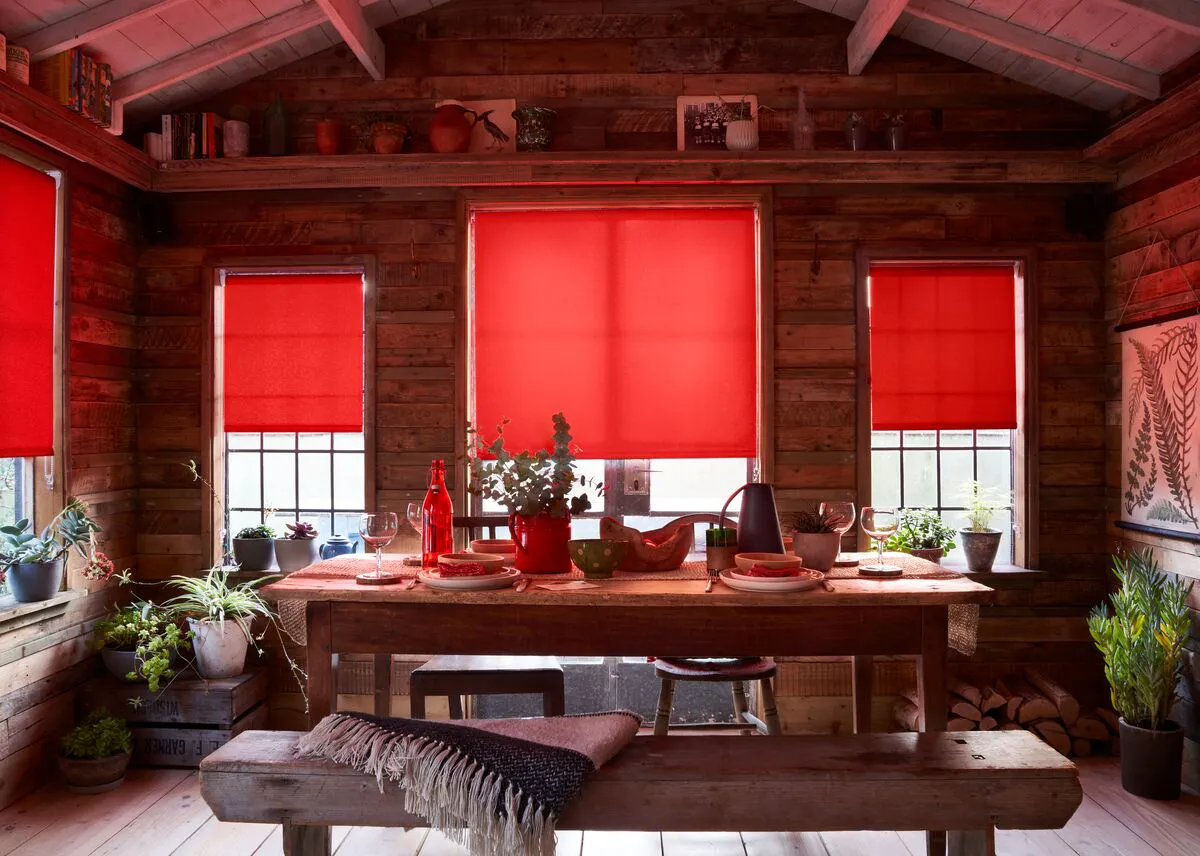 Rustic wooden dining room with red roller blinds, potted plants and a wooden table set with red-toned tableware.