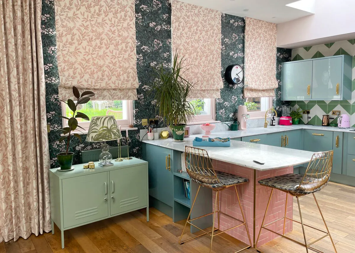 Colourful kitchen with patterned wallpaper, blush Roman blinds, pastel cabinets and a marble island with gold bar stools.