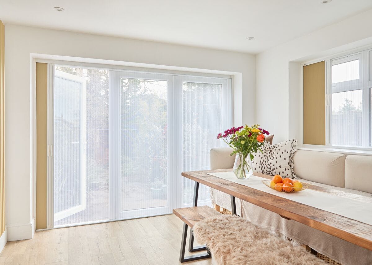 Bright dining nook with sheer white blinds on patio doors, a wooden table with flowers and fruit, and a cream corner bench with cushions.
