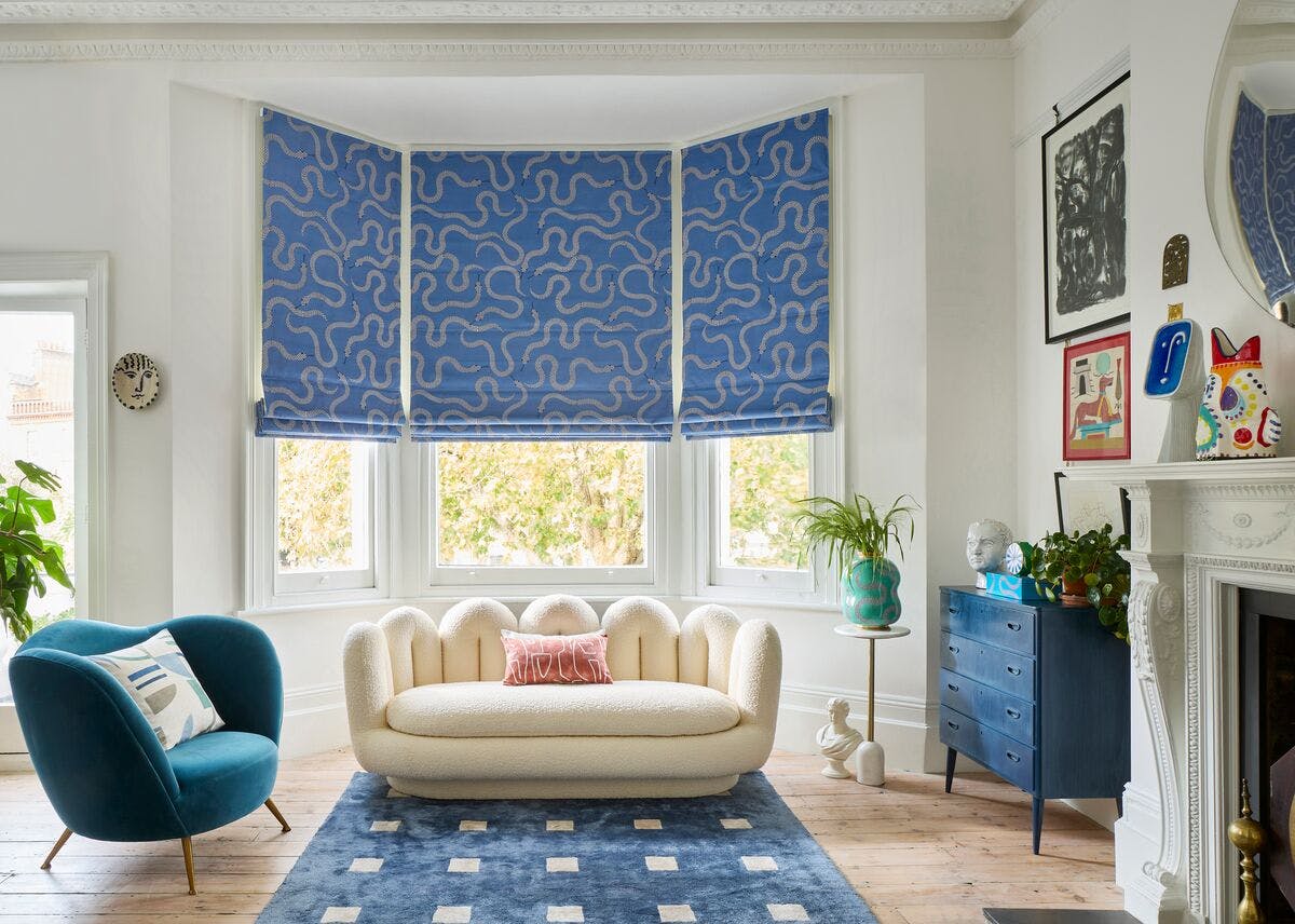 Bright living room with blue patterned Roman blinds, cream sofa, teal armchair and modern artwork around a large bay window.