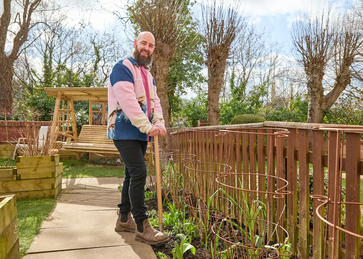 Person gardening beside a wooden fence on a sunny day, tending to plants along a garden path with trees and a swing bench in the background.