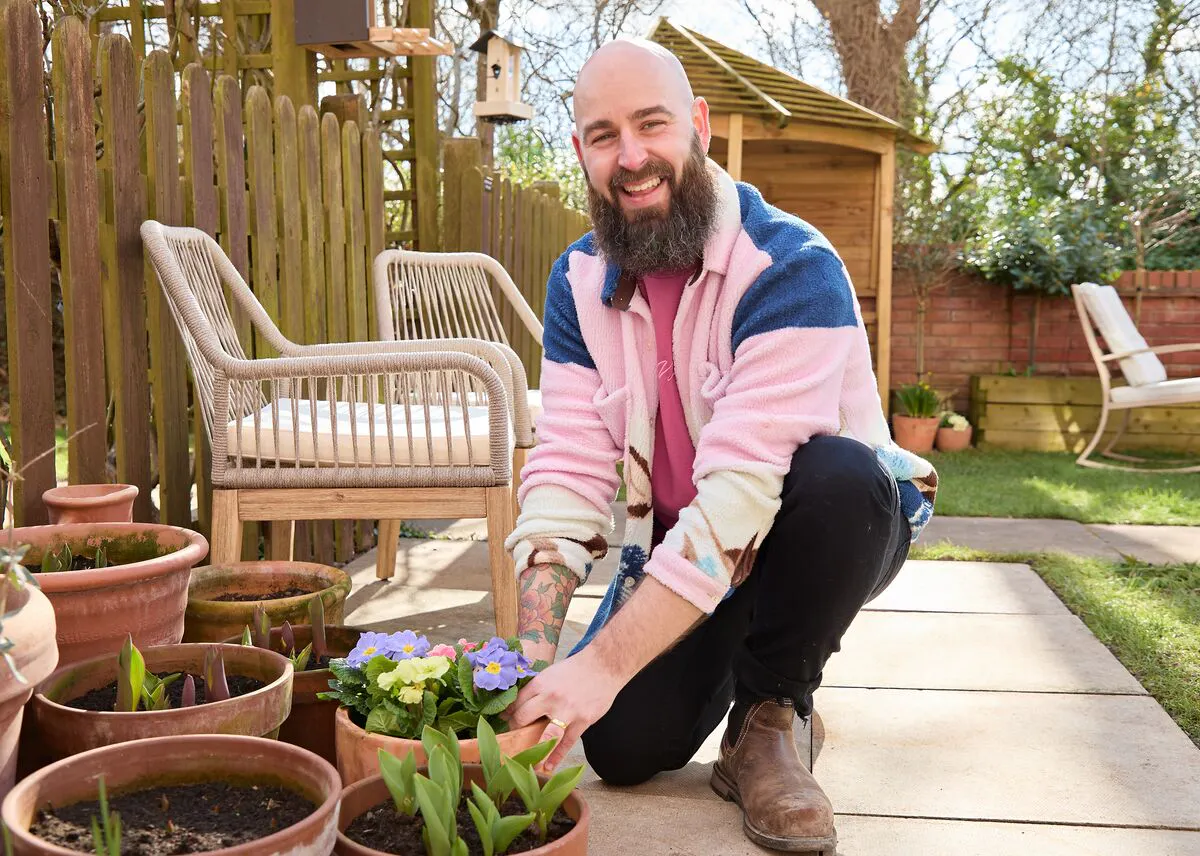 Influencer Luke Newnes, wearing a pink and blue fleece in his garden planting purple and yellow flowers in a pot with multiple pots and shoots around on a sandstone-style patio next to two raffia chairs with cream cushions.