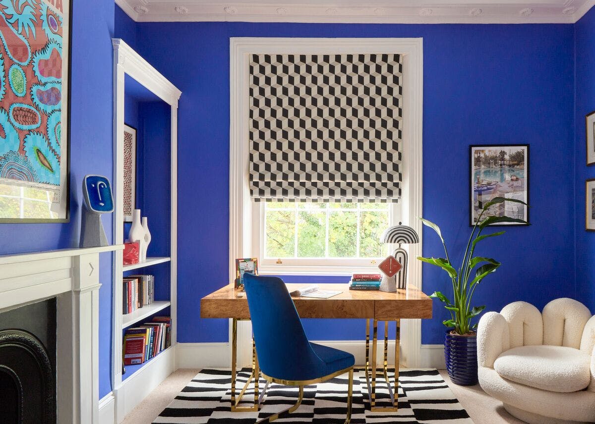 Bold blue home office with a geometric black‑and‑white Roman blind, wooden desk, blue chair and modern décor around a central window.