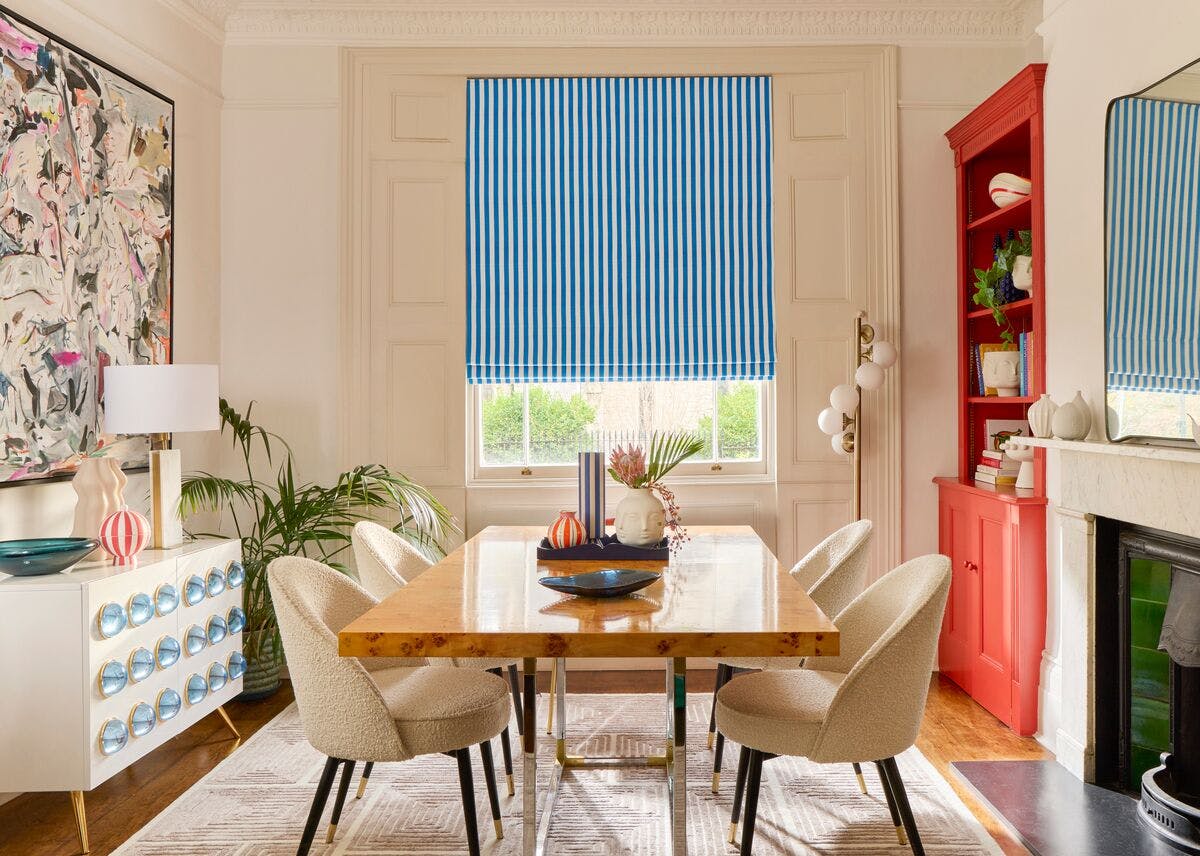 Modern dining room with a bright blue striped Roman blind, wooden dining table, cream chairs and bold colourful décor.