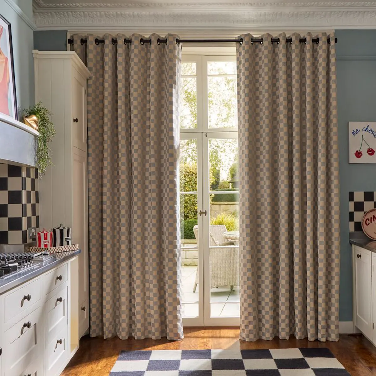 Classic kitchen with checkerboard curtains at French doors, cream cabinets, chequered tiles and a view to a sunlit patio garden.