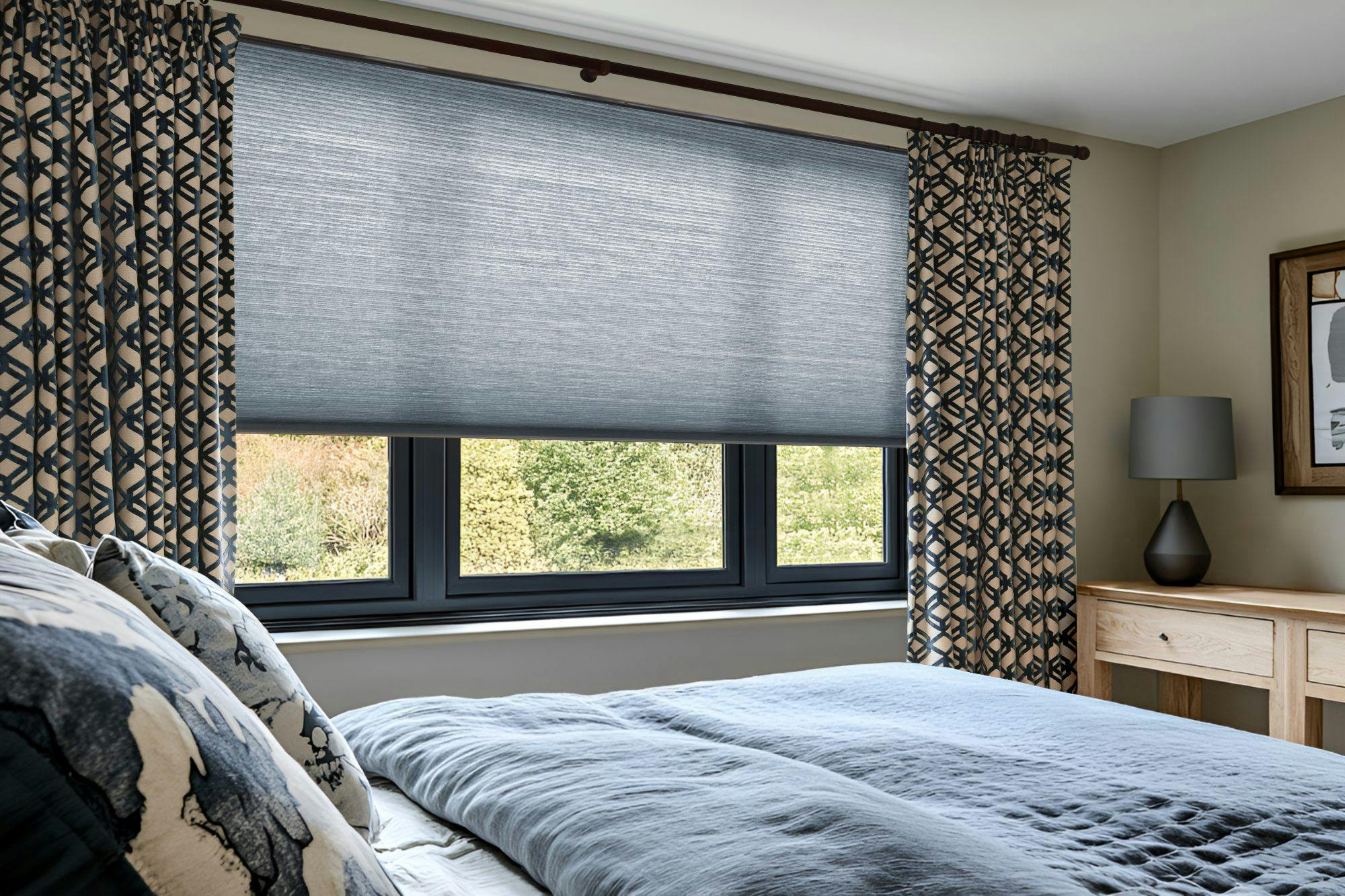 Modern bedroom with patterned curtains and a grey pleated window blind, featuring a wooden bedside table, decorative lamp, and a neatly made bed with blue bedding.