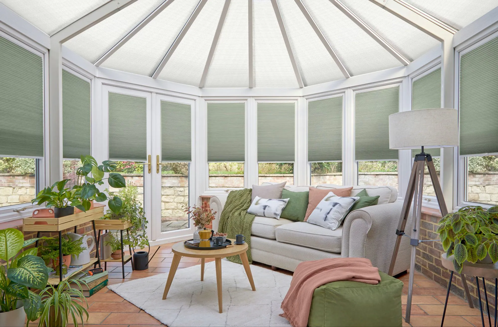 Large round conservatory with white frames dressed in forest green duette blinds to side, lowered halfway, and white duette roof blinds, closed, terracotta tiled floor, cream sofa with cushions in green, terracotta and white with grey touches, green square pouffe with terracotta throw, white rug, tripod lamp with white shade, lots of plants throughout.