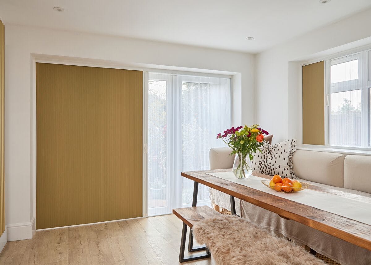 Bright dining nook with mustard sliding blinds, sheer door panels, a wooden table with bench seating and a vase of colourful flowers.