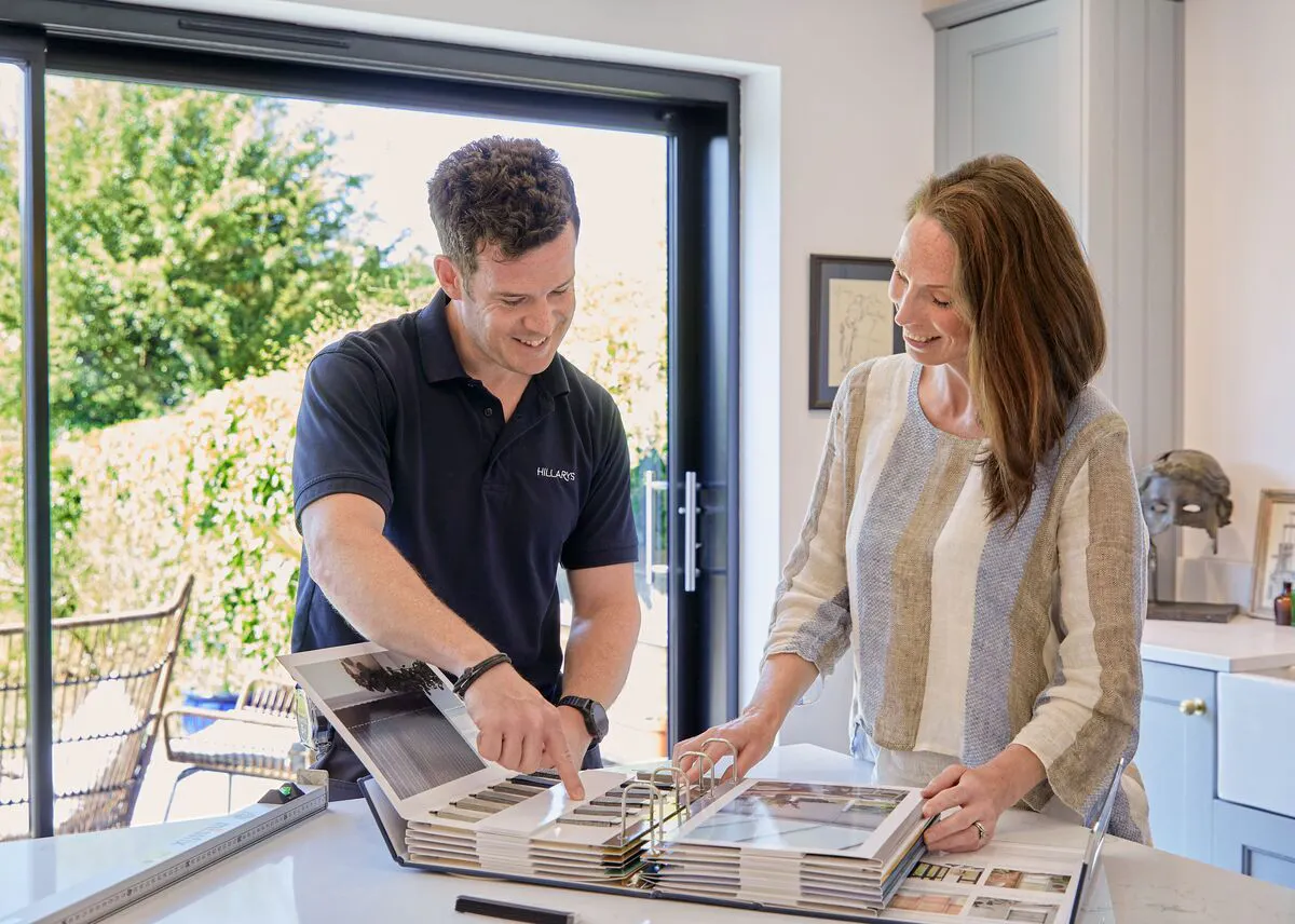 Two people reviewing fabric samples together at a kitchen table, with large patio doors behind them letting in natural light.