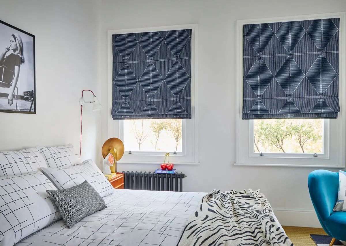 Bright bedroom with two navy geometric Roman blinds, monochrome grid bedding, a gold lamp on a wooden bedside table and a blue accent chair.