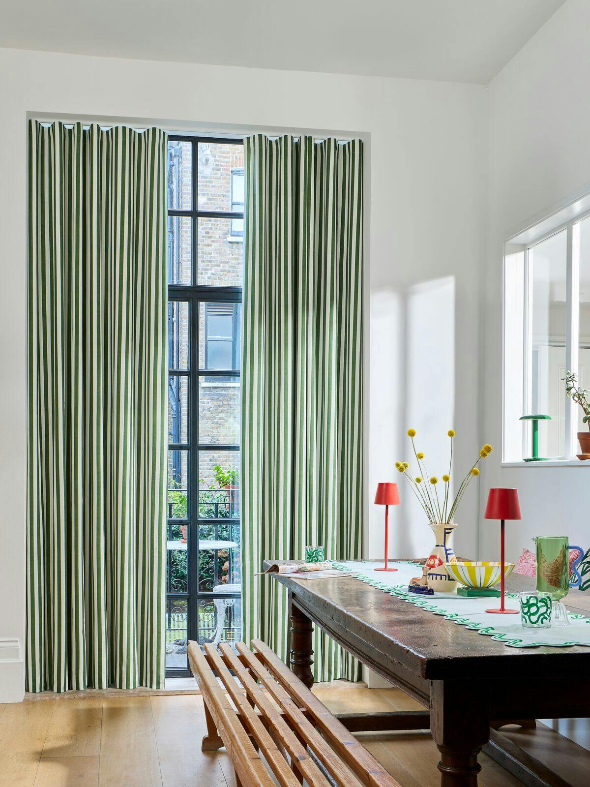 Bright dining room with green striped curtains, a wooden table and bench, and colourful decor arranged near tall black-framed windows.