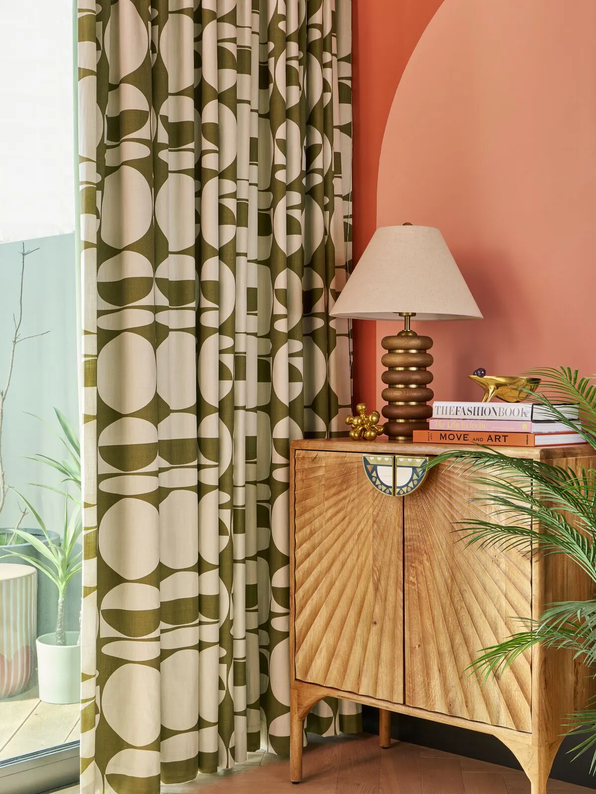Corner of a colourful room with green patterned curtains, a wooden cabinet, table lamp, books and indoor plants beside a large window.