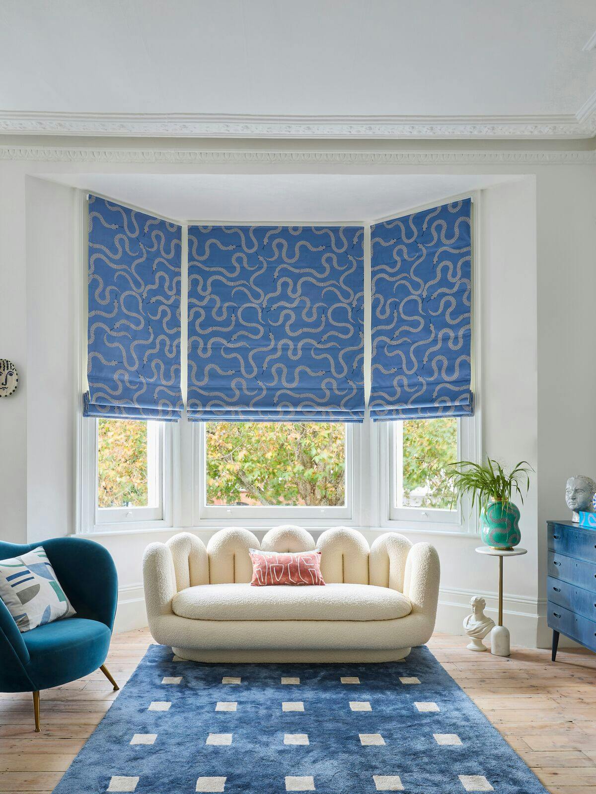 Bright living room with blue patterned Roman blinds, cream sofa, teal armchair and modern artwork around a large bay window.