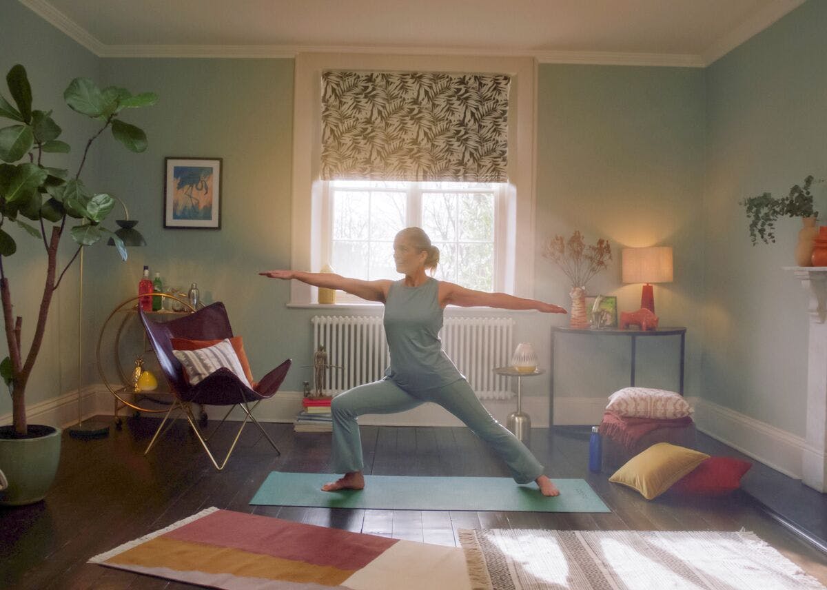 Yoga pose in a cosy living room with soft lighting, patterned blinds, plants, and cushions around a mat.