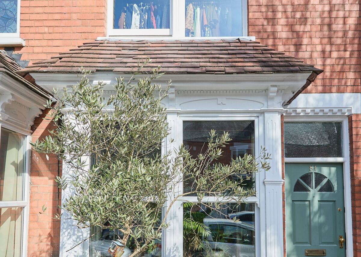 Front view of a brick house with a bay window, green front door, and a leafy tree in the garden.