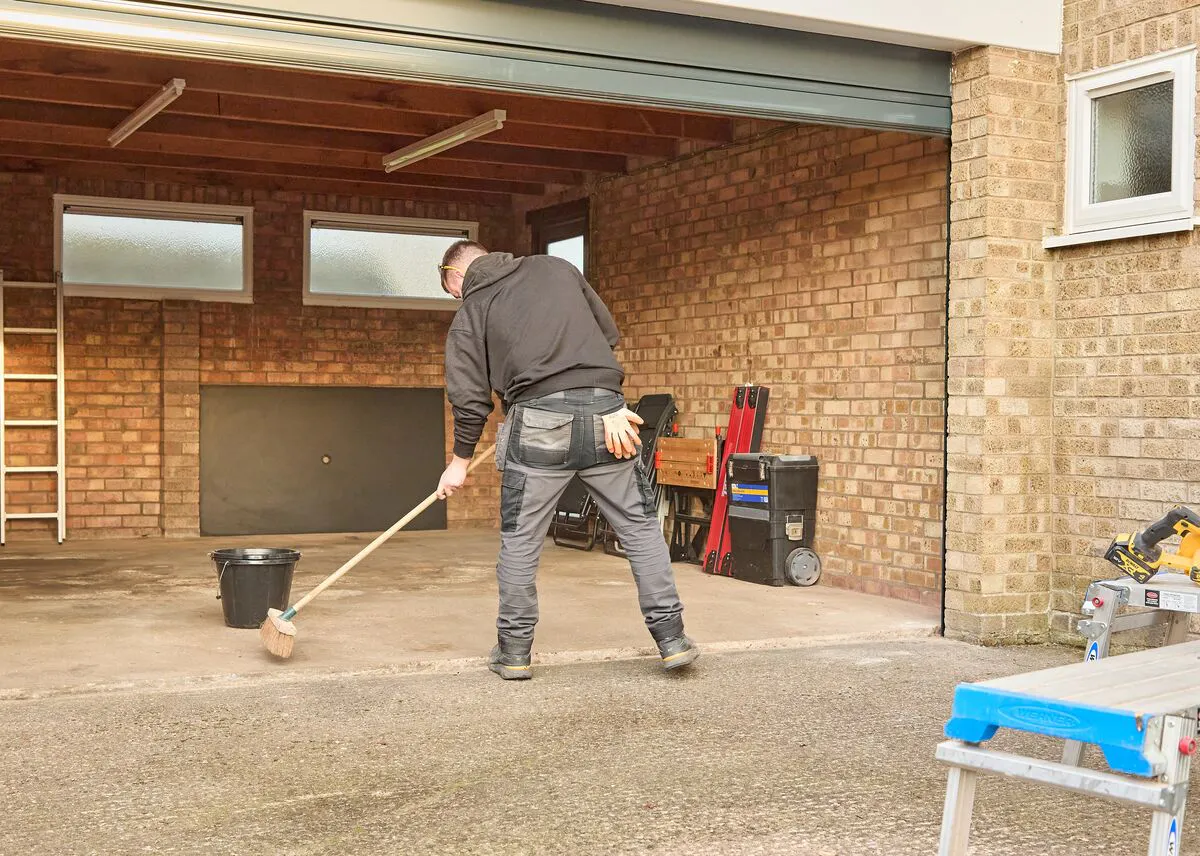 Worker sweeping the floor of an empty brick garage during a garage door installation.