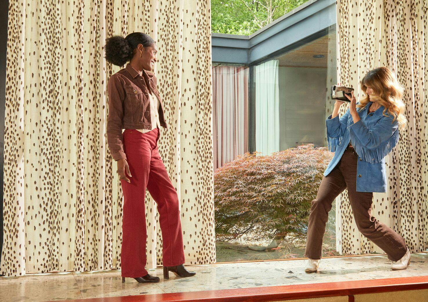 Fashion photoshoot scene with one person posing indoors while another person takes a picture with a camera, featuring patterned curtains and large windows with garden views, styled to showcase the Winnie and Kira Poodle and Blonde collection.