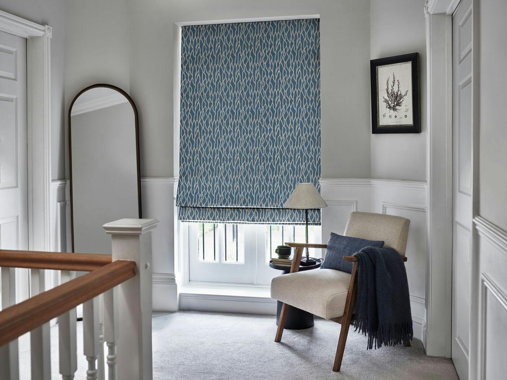 Calm, light-filled landing area with a blue patterned Roman blind, modern armchair with a throw, side table with a lamp, and a tall arched mirror.
