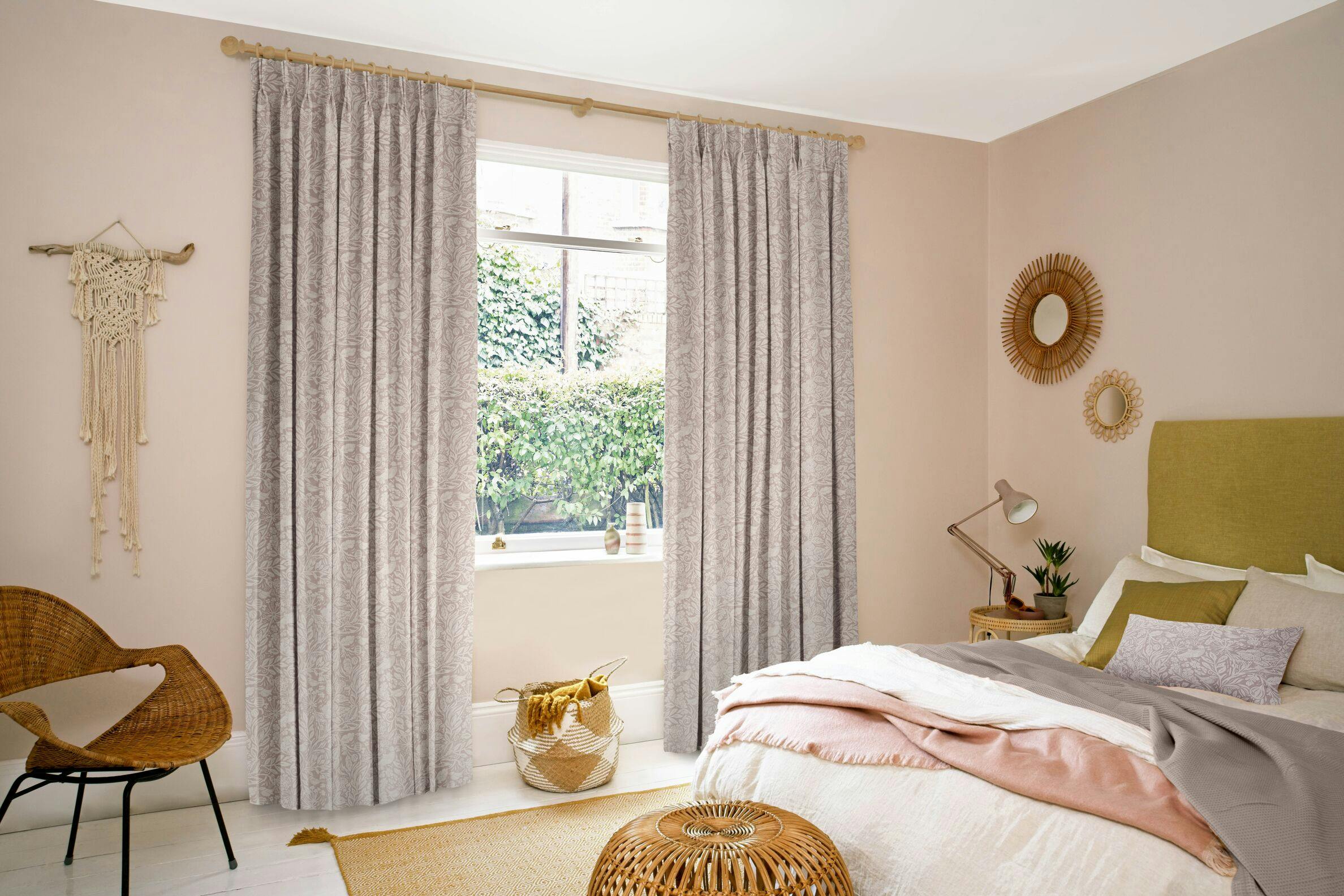 Lilac curtains hang beside a window adorned with greenery, in a cozy bedroom with a bed, wicker chair, and decorative items. Soft light creates a serene, inviting atmosphere.