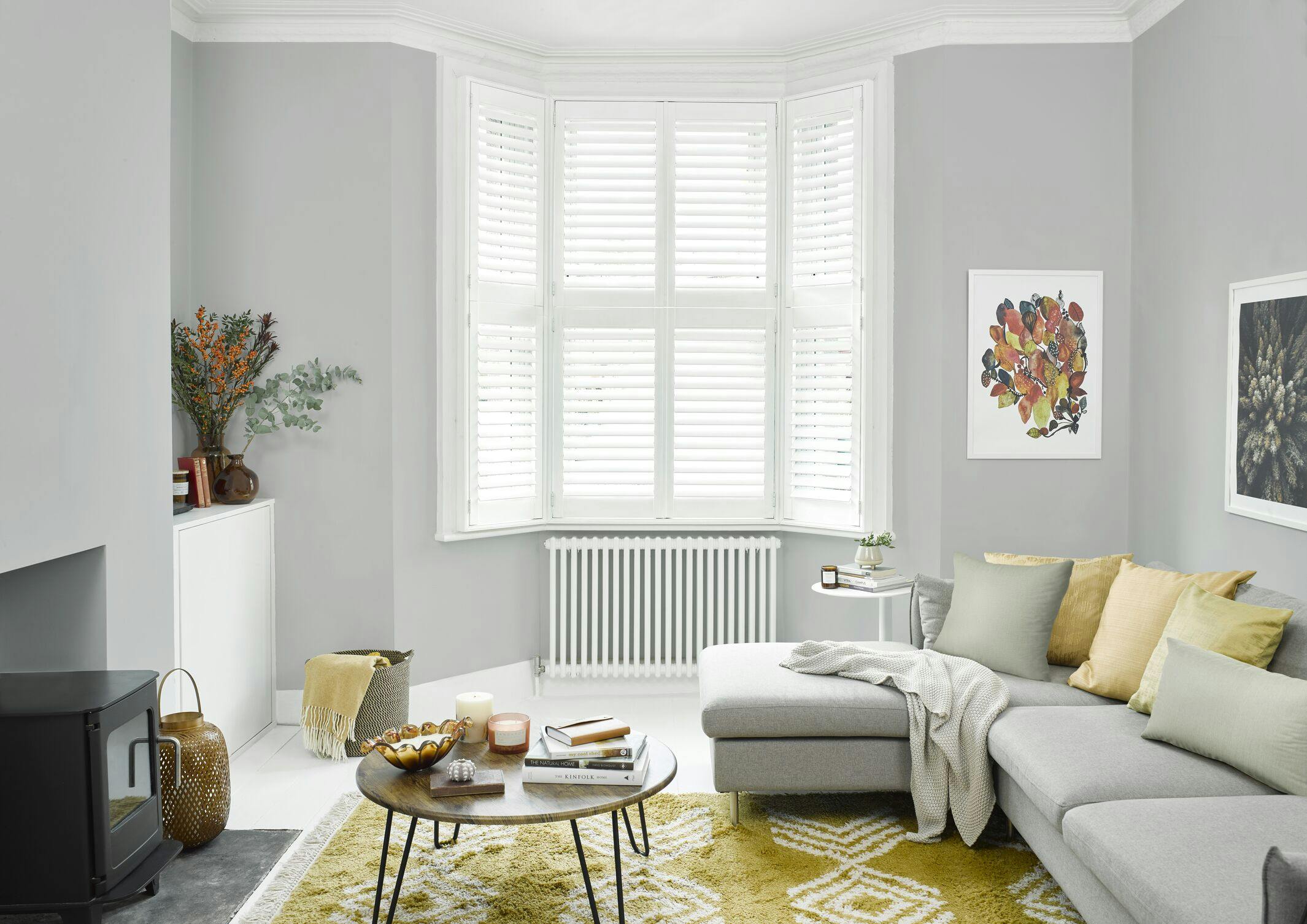 A cozy living room features a gray sectional sofa with patterned cushions. A wooden coffee table with books and candles sits on a yellow rug. White shutters cover bay windows.