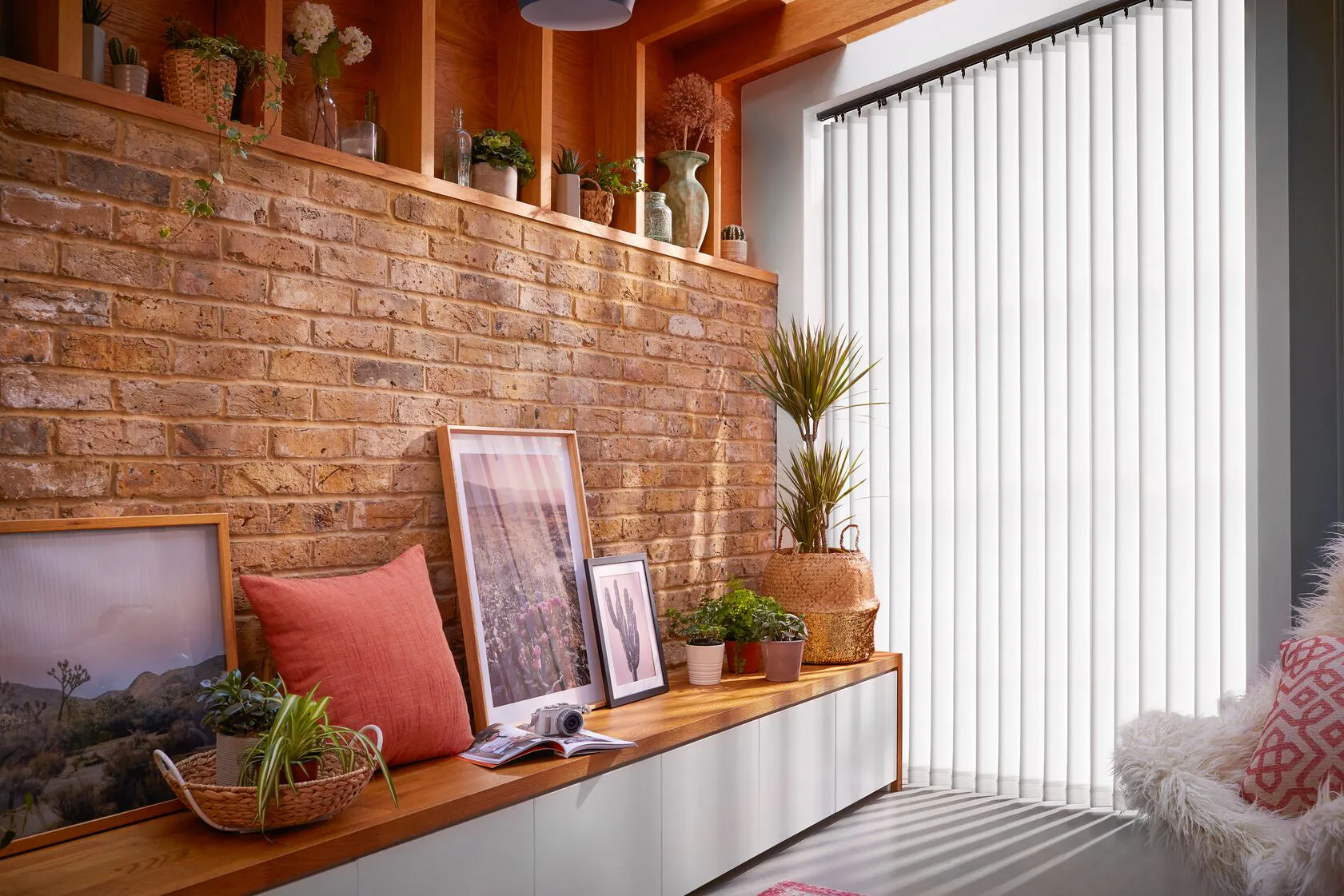 Framed photographs and a red pillow rest on a wooden bench against a brick wall. Various plants decorate the space. Vertical blinds cover a window, softly filtering daylight.