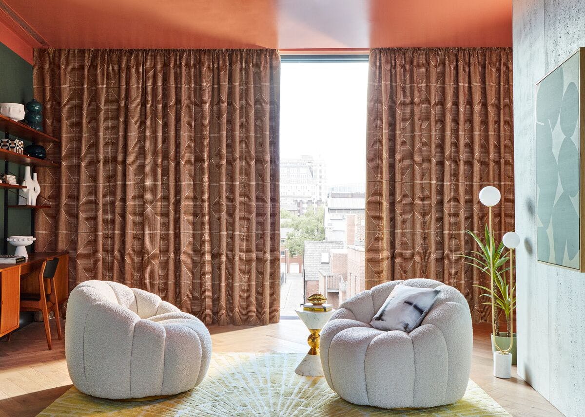 Cosy modern room with two curved armchairs, patterned floor‑to‑ceiling curtains, warm lighting, and a view of city buildings through a tall window.
