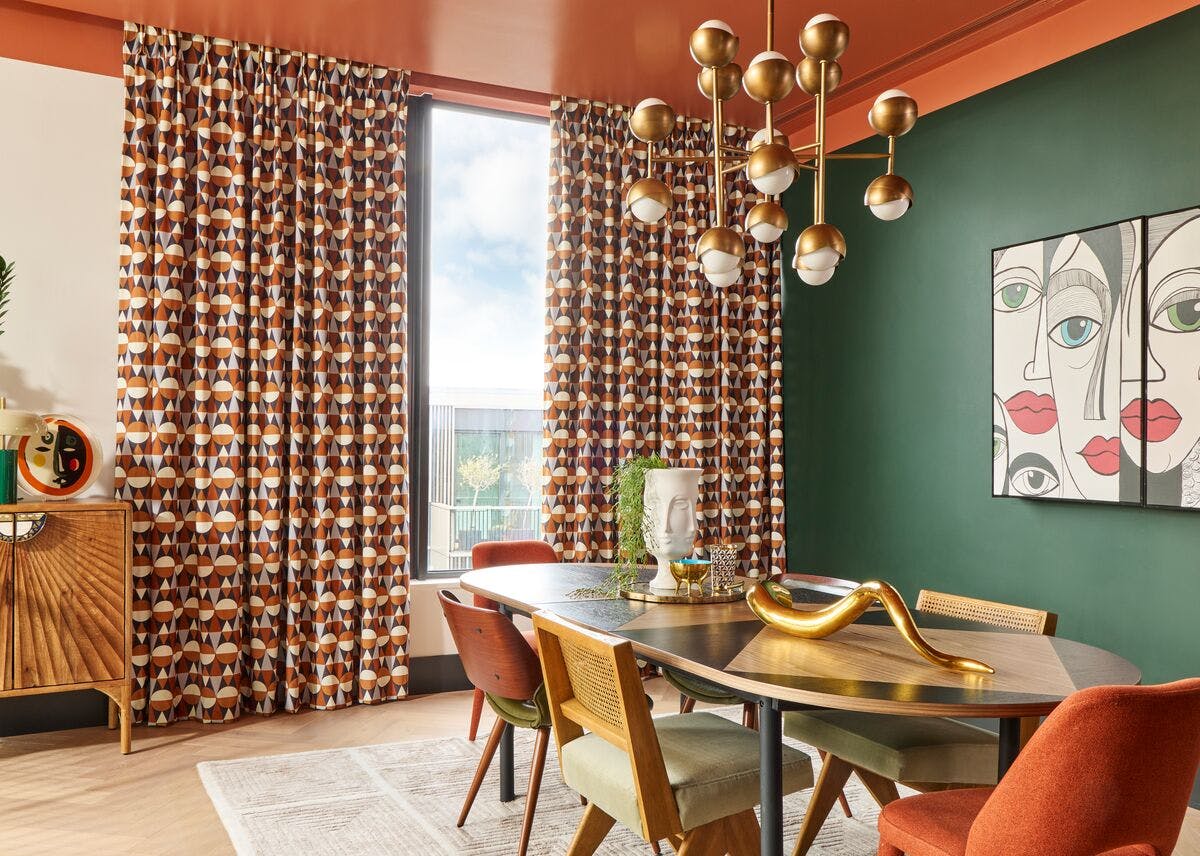 Colourful dining room with patterned curtains, a modern chandelier, green feature wall, and a wooden table with mixed retro‑style chairs.