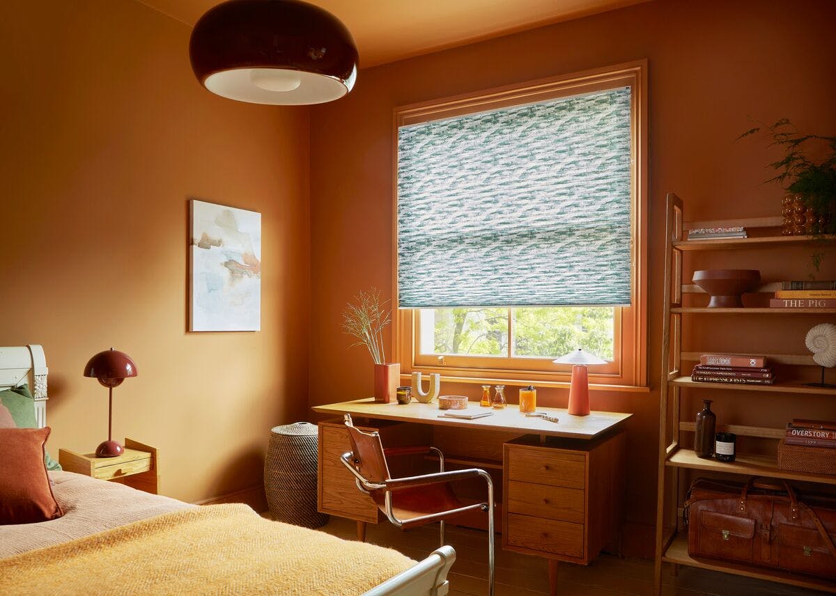 Warm bedroom‑study with a wooden desk, patterned Roman blind, shelving, and earthy décor in soft orange tones.