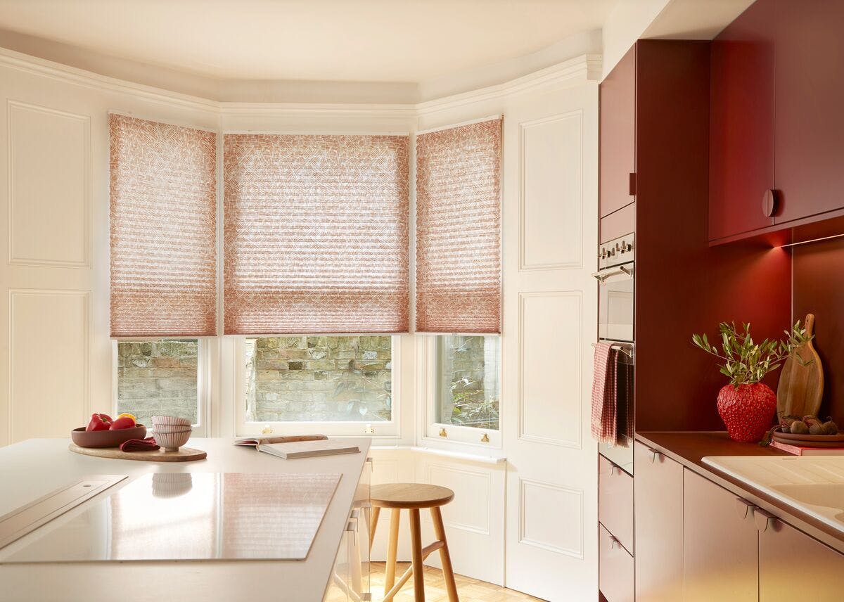 Bright kitchen with bay window featuring textured pleated blinds, white cabinetry, and warm red accents with modern worktops and decor.