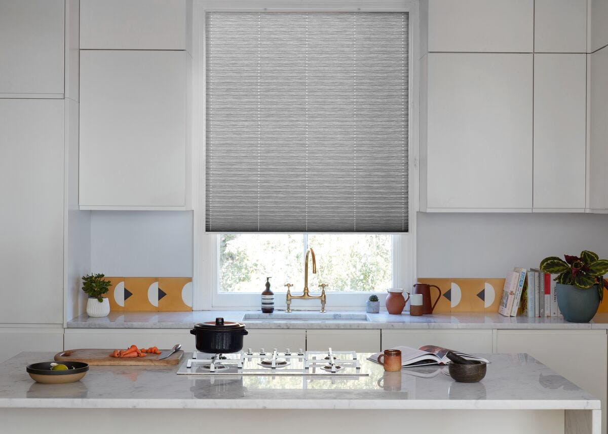 Bright modern kitchen with white cabinets, marble island, and a grey pleated blind over the window above a gold tap.