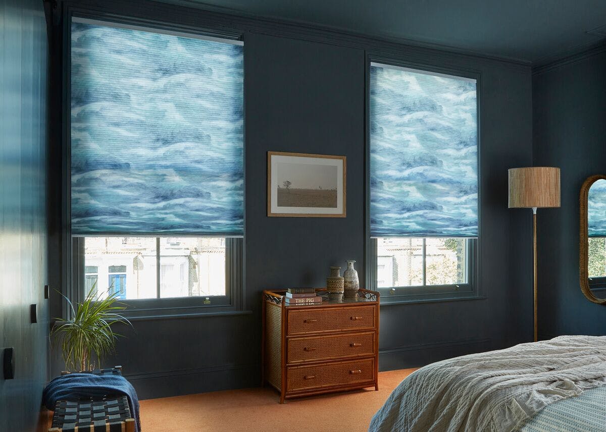 Calm blue bedroom with two windows featuring cloud‑patterned blinds, a wooden chest of drawers, plants, and a neatly made bed.