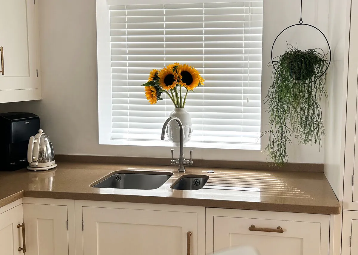 Kitchen sink beneath white venetian blinds, vase of sunflowers on the ledge, beige worktop, kettle and a hanging plant adding greenery.