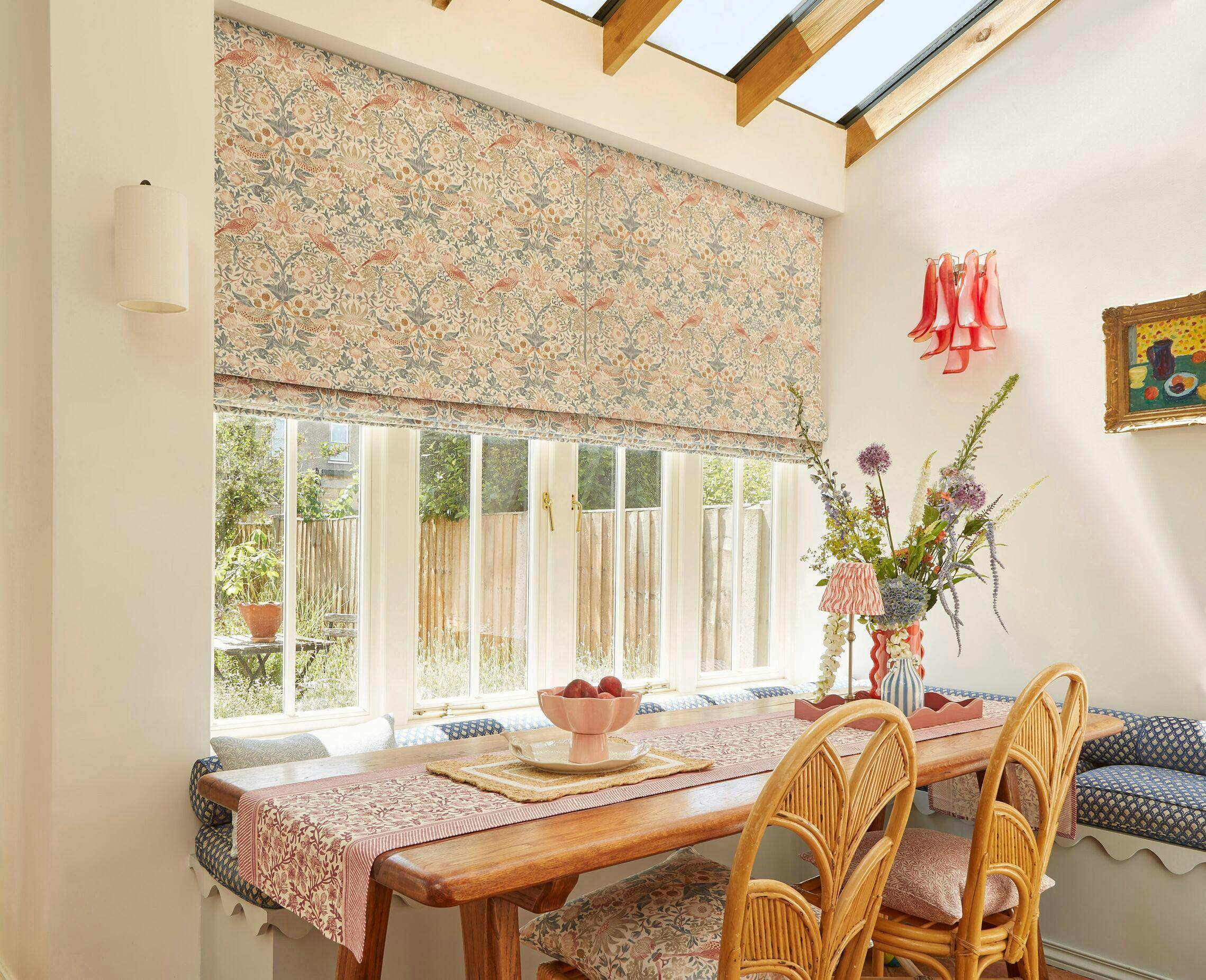 Close up corner of a room with windows on one side dressed in two Roman blinds with pale pink, salmon and blue pattern, lowered halfway, windows in sloped roof above, pale wooden table with pink patterned runner and two chairs, banquette seating in L-shape with blue and white polka dot cushions.