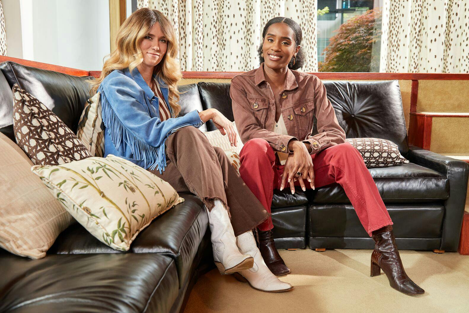 Two people sit on a black leather couch, smiling, in a warmly lit room with decorative pillows and patterned curtains. One wears a blue jacket, the other a brown jacket.