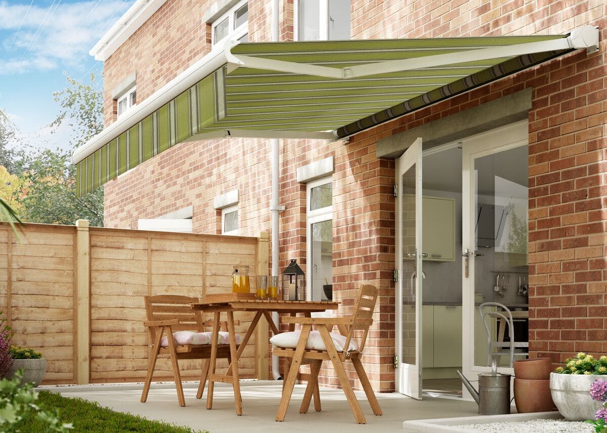 A green retractable awning shades a patio with wooden furniture outside a brick house, near a green fence and garden plants.