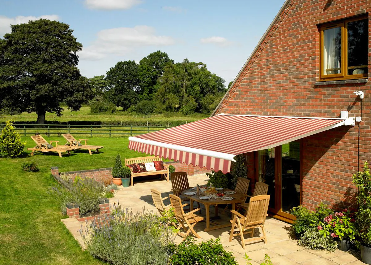 Patio with wooden dining set beneath a red‑striped awning, surrounded by a sunny garden with loungers, plants and open countryside.