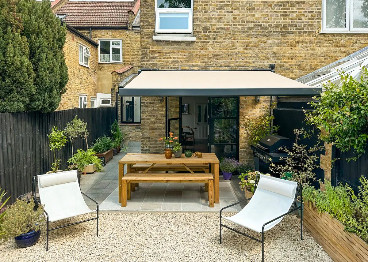 Small garden patio with a beige awning, wooden dining table, lounge chairs and plants against the brick exterior of a house.