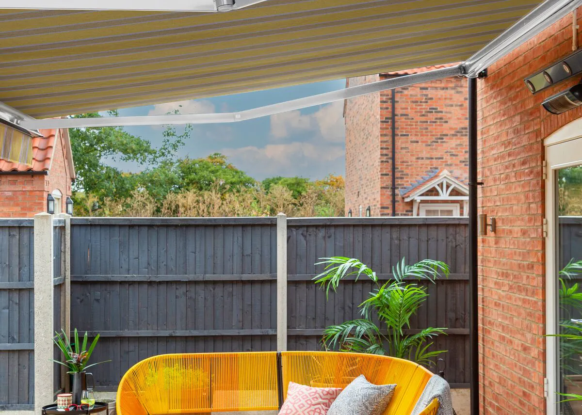Patio seating area with a yellow striped awning, bright yellow outdoor sofa, potted plants and a view of a garden beyond a grey fence.