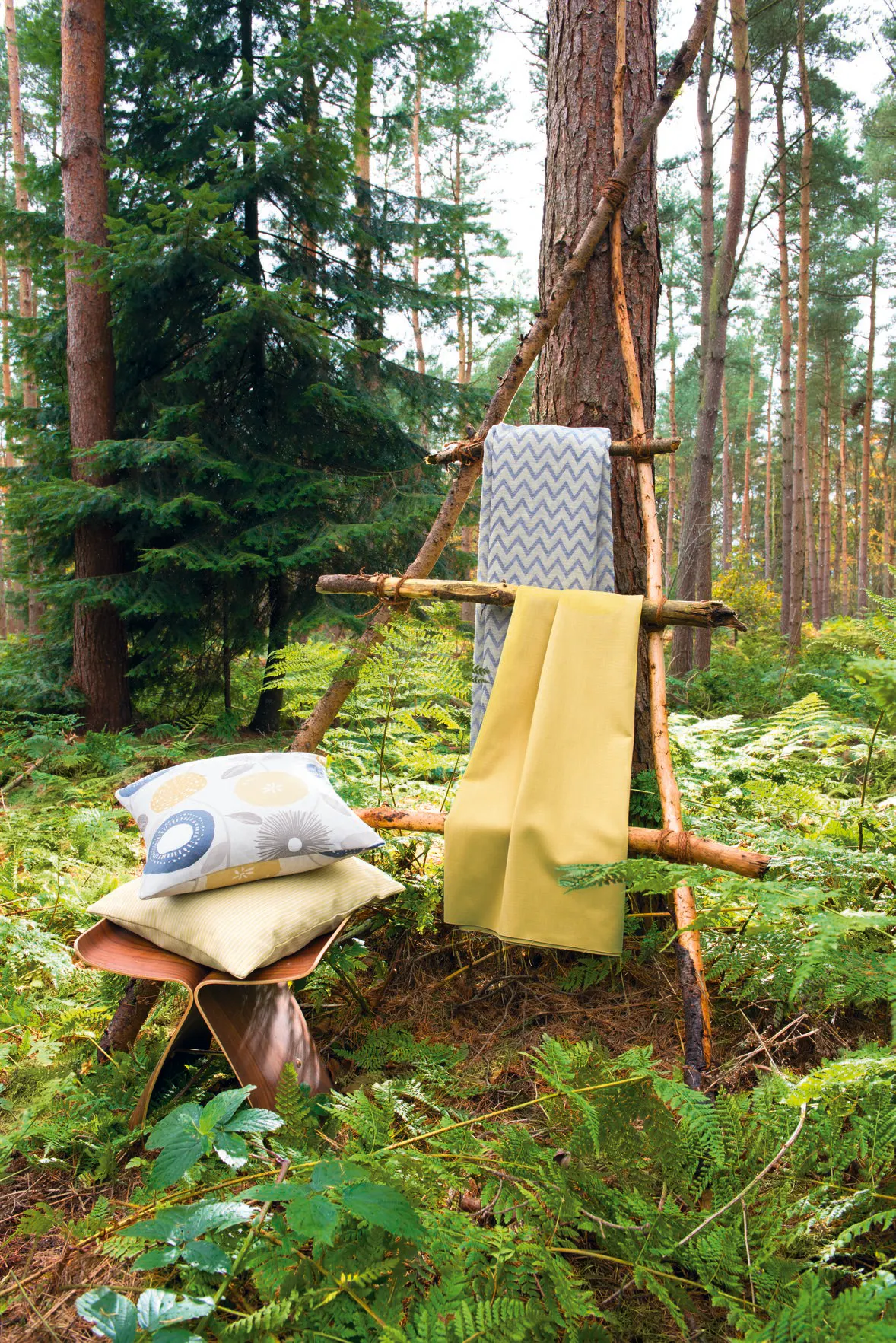 Fabrics and patterned cushions displayed on a rustic wooden frame in a lush forest setting with ferns and tall pine trees.