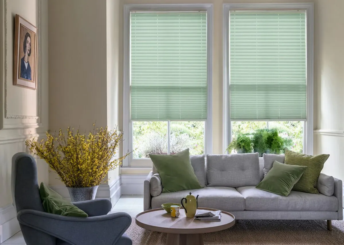 Bright living room with light green pleated blinds, grey sofa with green cushions, armchair, and yellow flowers.