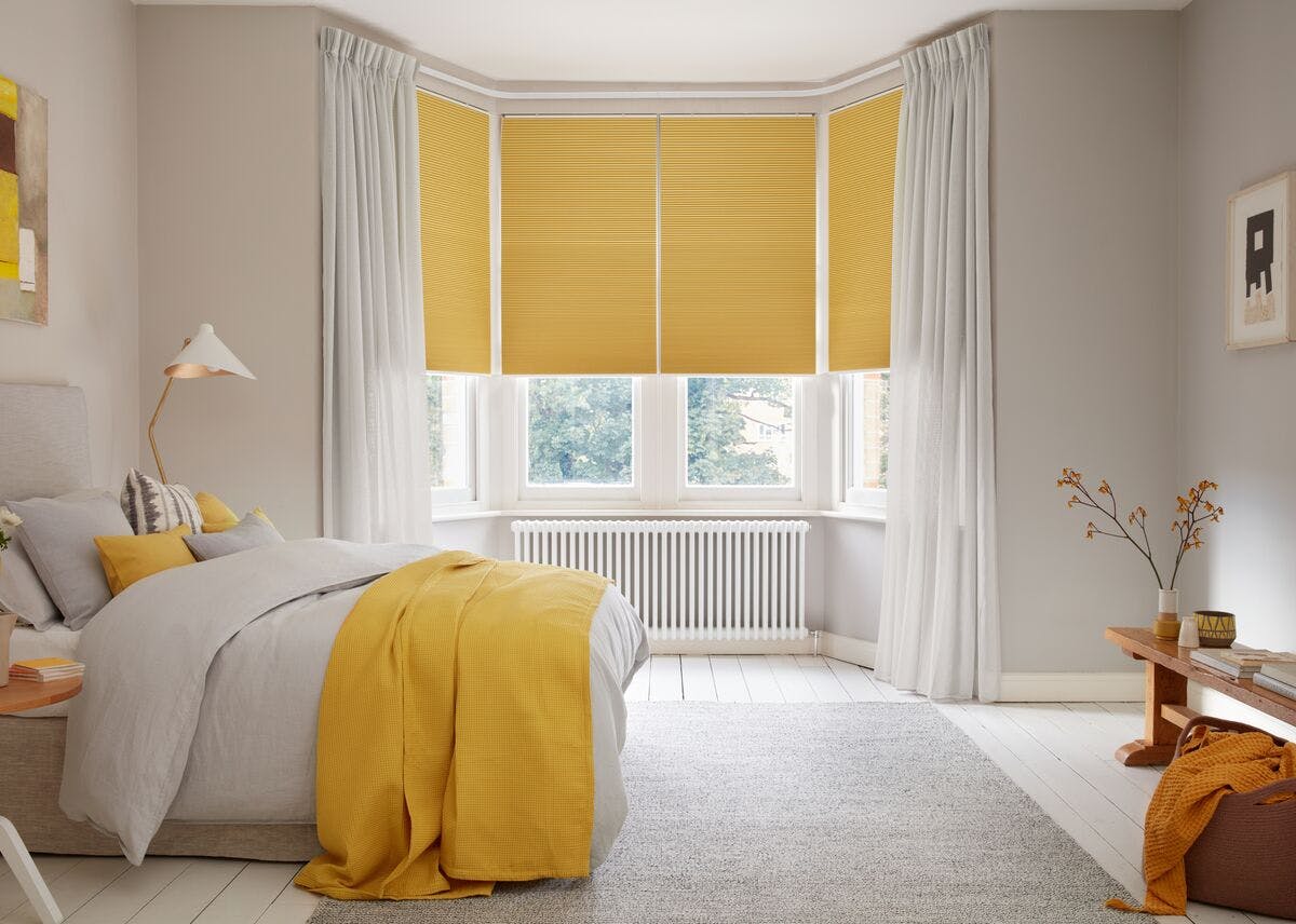 Cream bedroom with bay window dressed in a sunshine yellow honeycomb blind, lowered halfway, with sheer white voiles open to the sides, bed with white bedding with sunshine yellow throw and cushions, wooden bench, modern art in yellow and beige.